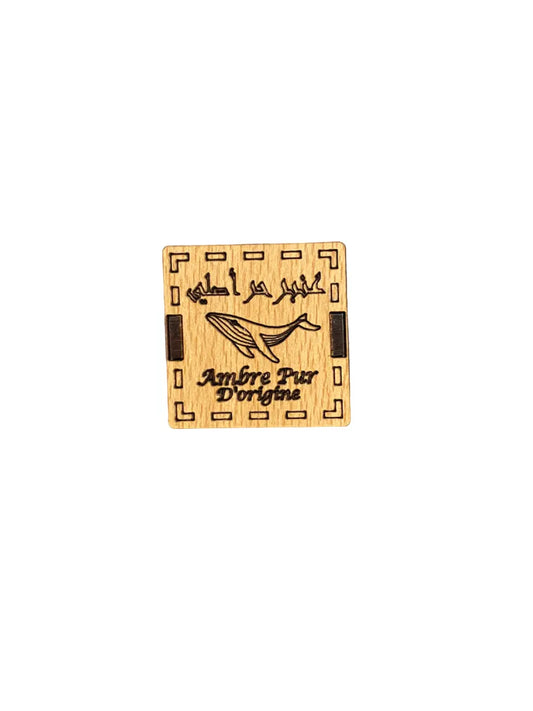 Ambre de mer - Ambar el bahar wooden label featuring Arabic calligraphy, a decorative whale illustration, and geometric border pattern, showcasing authentic sea amber packaging design on light beige wood background