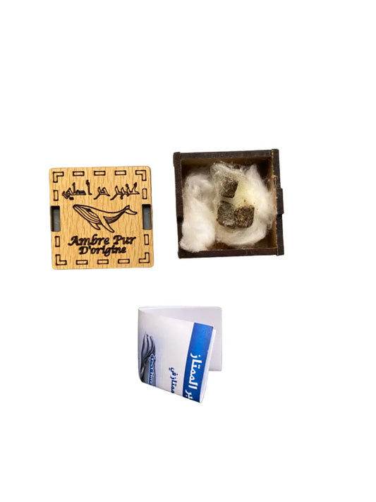 A piece of Ambre de mer (Ambar el bahar) displayed in a dark wooden box with cotton padding, alongside a decorative wooden cover featuring Arabic text and whale design, and product documentation with blue accents.